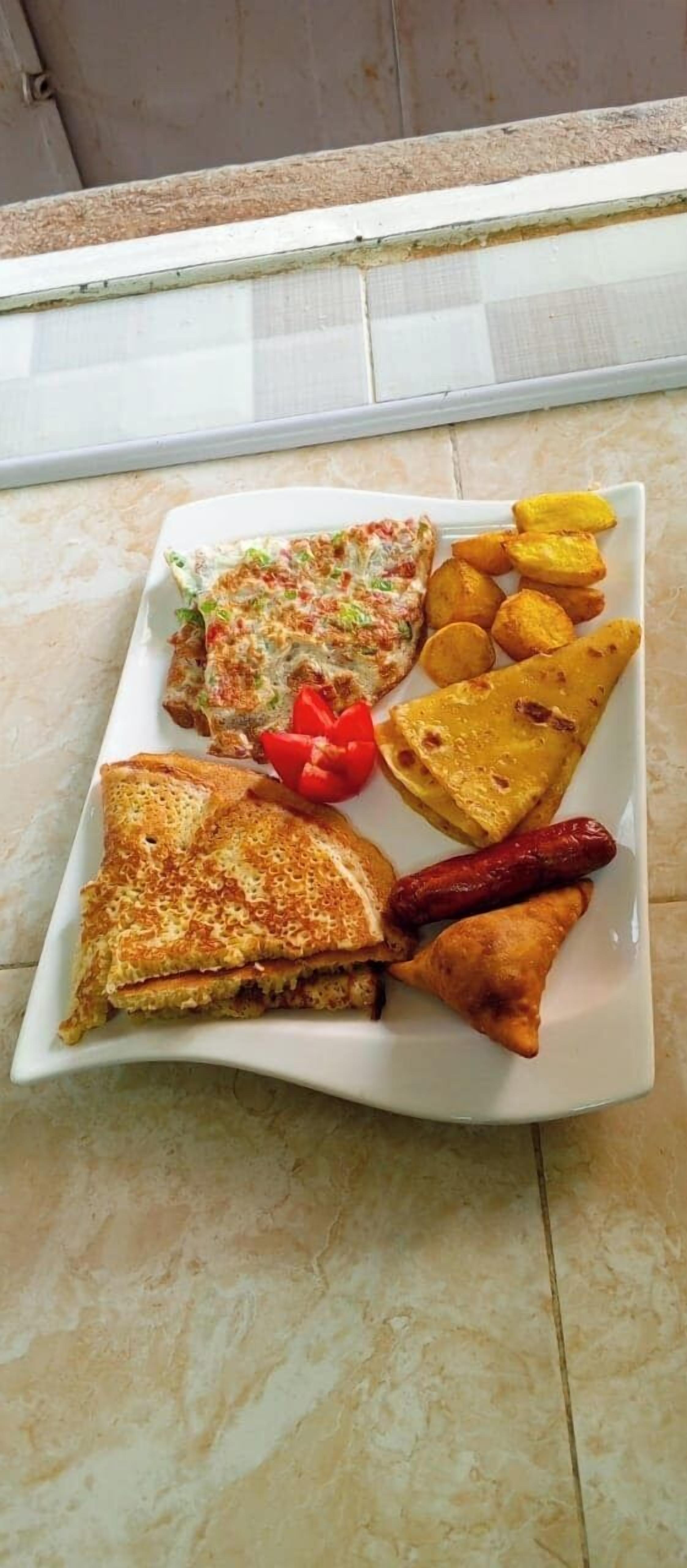 A square plate with a grilled sandwich, fried plantains, a red bell pepper, and a sausage.