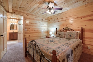 A wooden-paneled bedroom with a bed, a nightstand, a ceiling fan, and a bathroom visible through an open door.