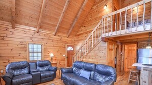A log cabin interior with two leather sofas, a dining area, and a staircase to an upper level.