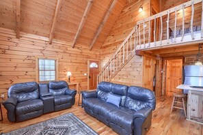 Ein Holzblockhaus-Interieur mit zwei Ledersofas, einem Essbereich und einer Treppe zu einer oberen Ebene.