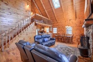 A log cabin interior with a wooden staircase, a leather sofa, a dining area, and a stone fireplace. - Woods Edge Cabin | Hocking Hills, Hot Tub, Pets (Logan)
