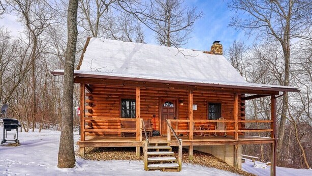 A log cabin with a snow-covered roof, wooden porch, and a chimney.