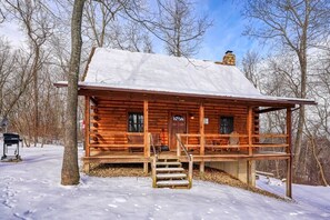 A log cabin with a snow-covered roof, wooden porch, and a chimney.