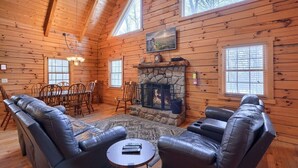 A cozy log cabin interior with a stone fireplace, leather sofas, a dining table, and large windows.