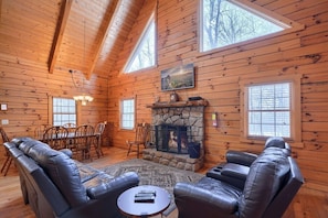 A cozy log cabin interior with a stone fireplace, leather sofas, a dining table, and large windows. - Woods Edge Cabin | Hocking Hills, Hot Tub, Pets (Logan)