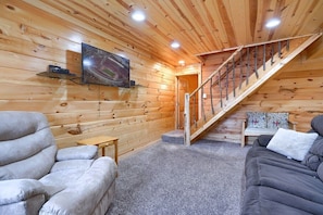 A log cabin interior with a wooden staircase, a sofa, a chair, and a mounted television. - Woods Edge Cabin | Hocking Hills, Hot Tub, Pets (Logan)