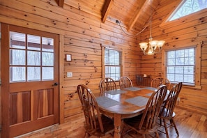 A wooden cabin interior with a dining table and chairs, wooden walls, and a chandelier.