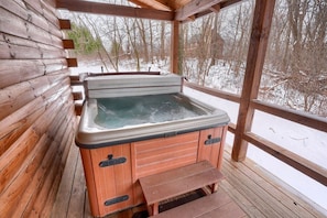 A wooden cabin with a hot tub on a wooden deck, surrounded by snow-covered trees.
