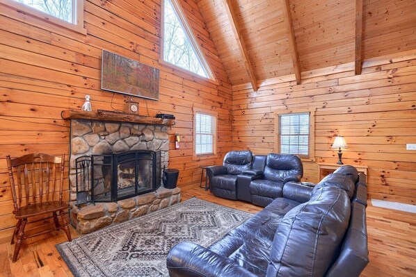 A log cabin interior with a stone fireplace, leather sofa, and a large window. - Woods Edge Cabin | Hocking Hills, Hot Tub, Pets (Logan)