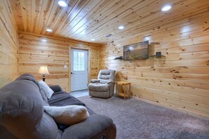 A cozy log cabin room with a sofa, a chair, a lamp, and a TV mounted on the wall.
