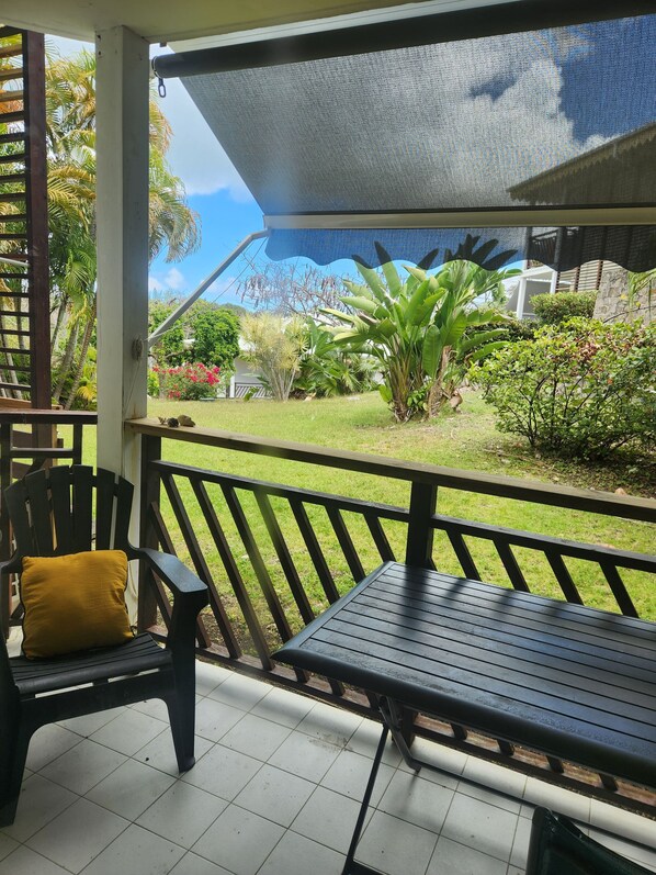 A covered outdoor seating area with a table and chairs, a view of a garden with various plants and a house in the background.