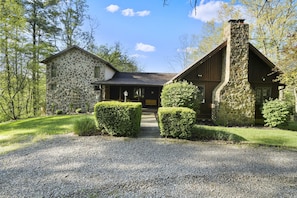 A stone house with a stone chimney, surrounded by a gravel driveway and trimmed hedges.
