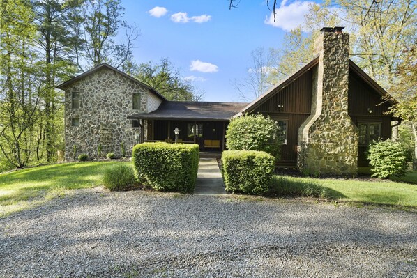 Une maison en pierre avec une cheminée en pierre, entourée d’une allée gravillonnée et de haies taillées.