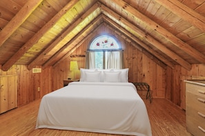 A wooden attic bedroom with a large bed, a window with curtains, and a wooden ceiling.