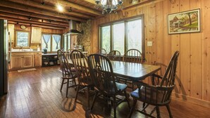 A wooden interior with a dining table and chairs, a kitchen area, and a large window with a view of trees.