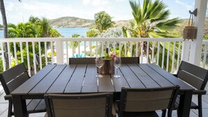 Une table en bois, entourée de quatre chaises, se trouve sur un balcon doté d  ’une balustrade blanche, d’où l’on peut admirer une plage et des collines.