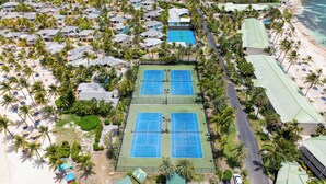Vue aérienne d’une station balnéaire comprenant plusieurs bâtiments de style bungalow, un court de tennis central et une plage aménagée avec des parasols.