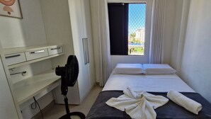 A small, clean room with a bed, a desk with drawers, a standing fan, and a window with curtains.