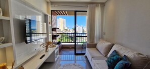 A modern living room with a flat-screen TV, a sofa, and a balcony with a city view. - CORVE0106 - Condomínio Villa di Capri (Salvador)