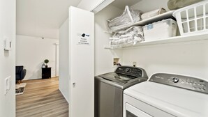 Une buanderie équipée d’un lave-linge et d’un sèche-linge, d’une étagère où le linge propre est plié, et d’un panneau indiquant la fonction de la pièce.