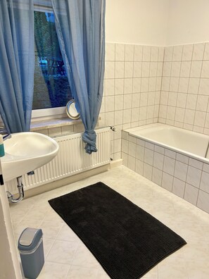A bathroom with a white sink, a bathtub, a window with blue curtains, and a black rug on the floor. - Cozy and quiet apartment in Rosenthal am Rennsteig (OT Blankenstein) (Rosenthal am Rennsteig)