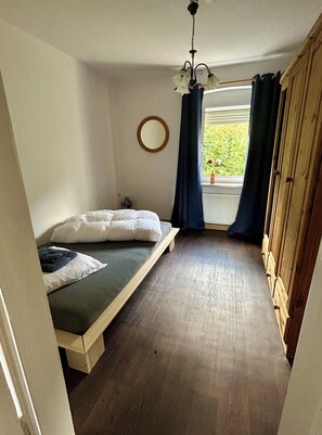 A bedroom with a single bed, a wooden wardrobe, a window with curtains, and a ceiling light fixture.