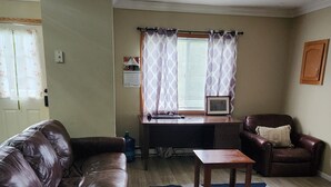 Ett vardagsrum med en brun lädersoffa, ett träbord och en brun läsfåtölj. Det står ett litet bord med ett tangentbord och en datorskärm på. Rummet har trägolv och ett fönster med mönstrade gardiner.