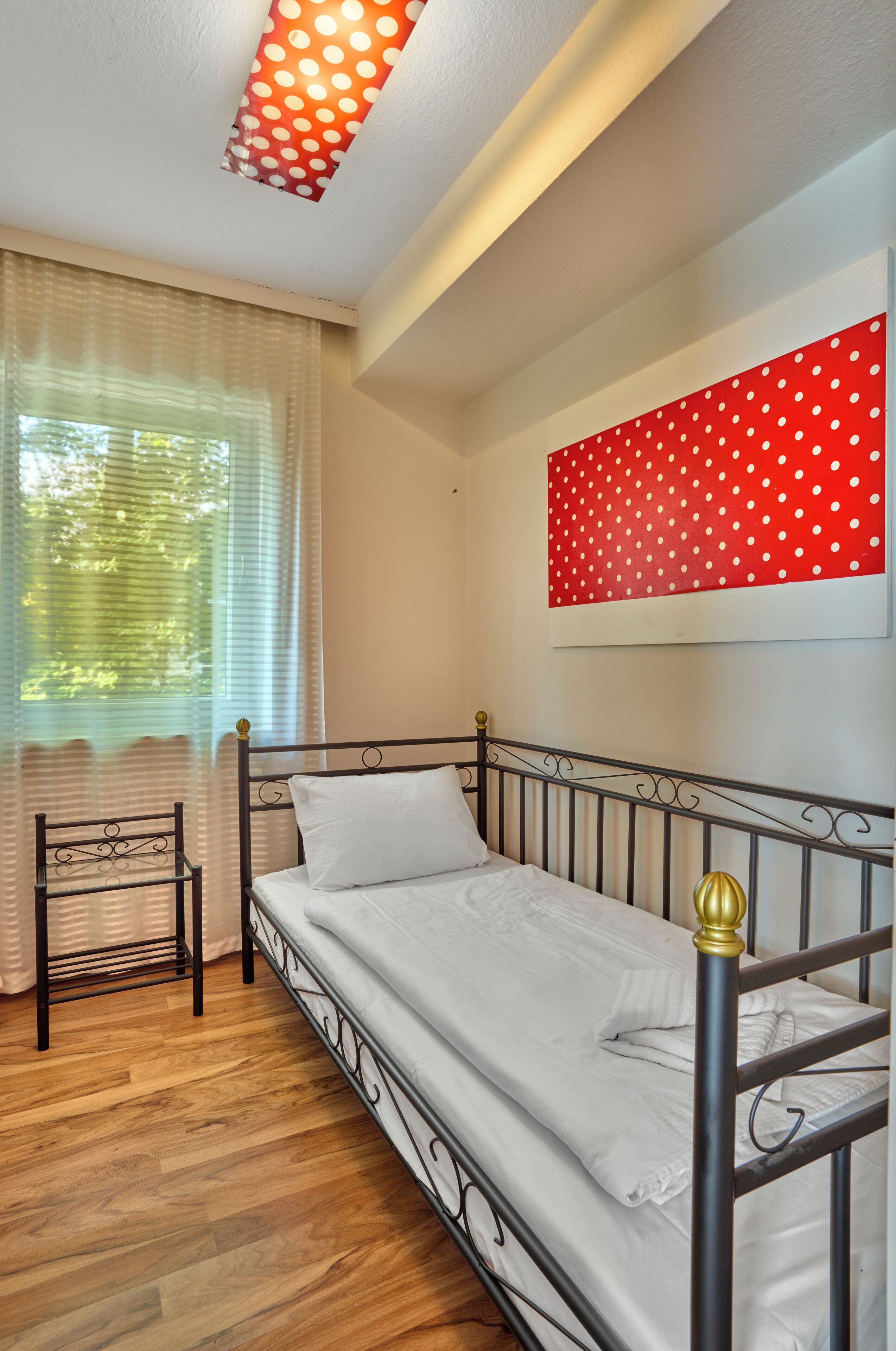 Un lit simple avec un cadre en métal et un matelas blanc, dans une pièce dont le sol est recouvert de parquet.