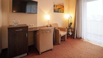 Habitación de hotel con televisión de pantalla plana, un escritorio con lámpara, dos sillas, una mesita con un florero y un ventanal grande con cortinas.