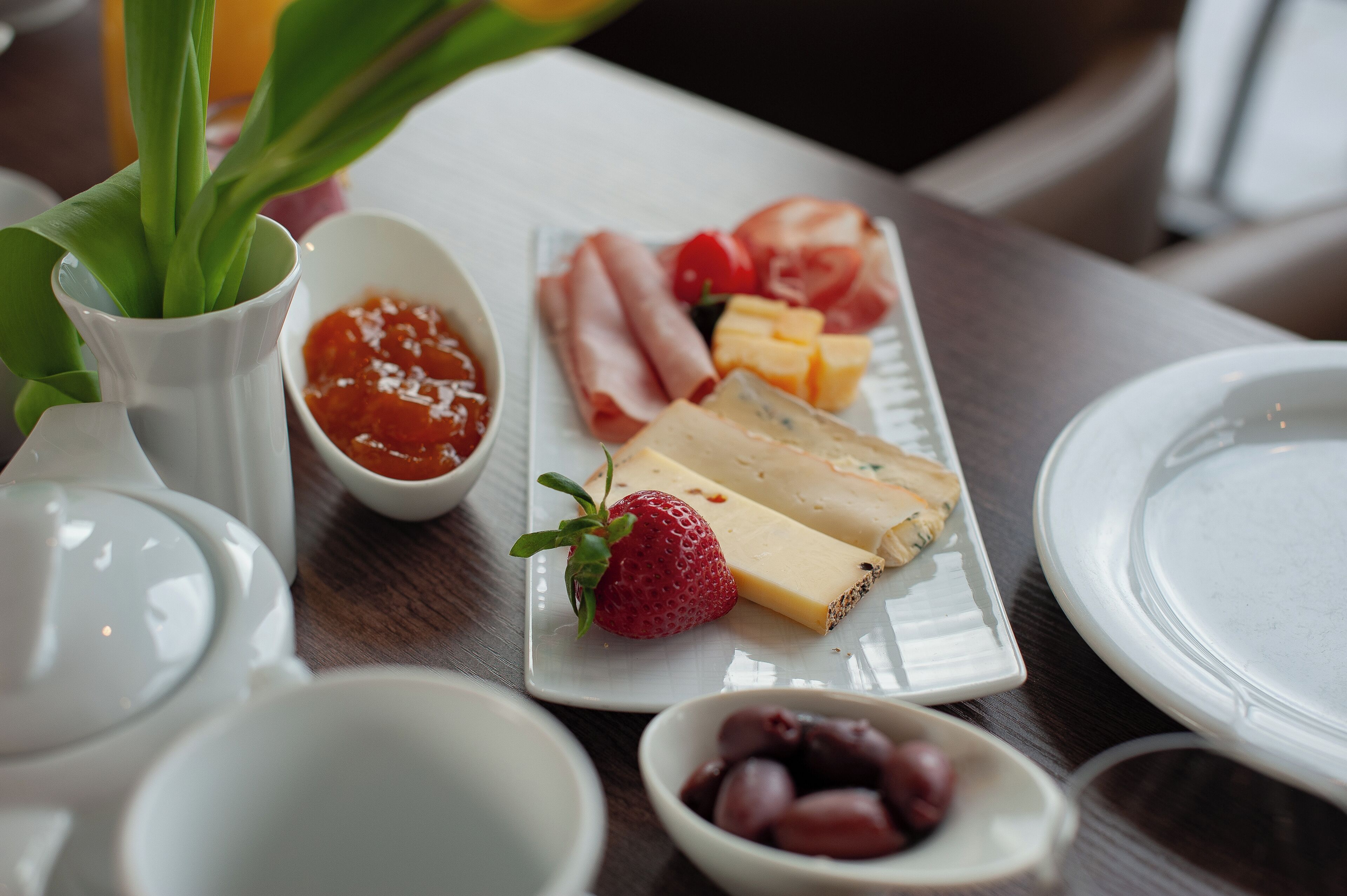 A table with a plate of cheese, strawberries, and ham, a bowl of jam, and a small bowl of olives.