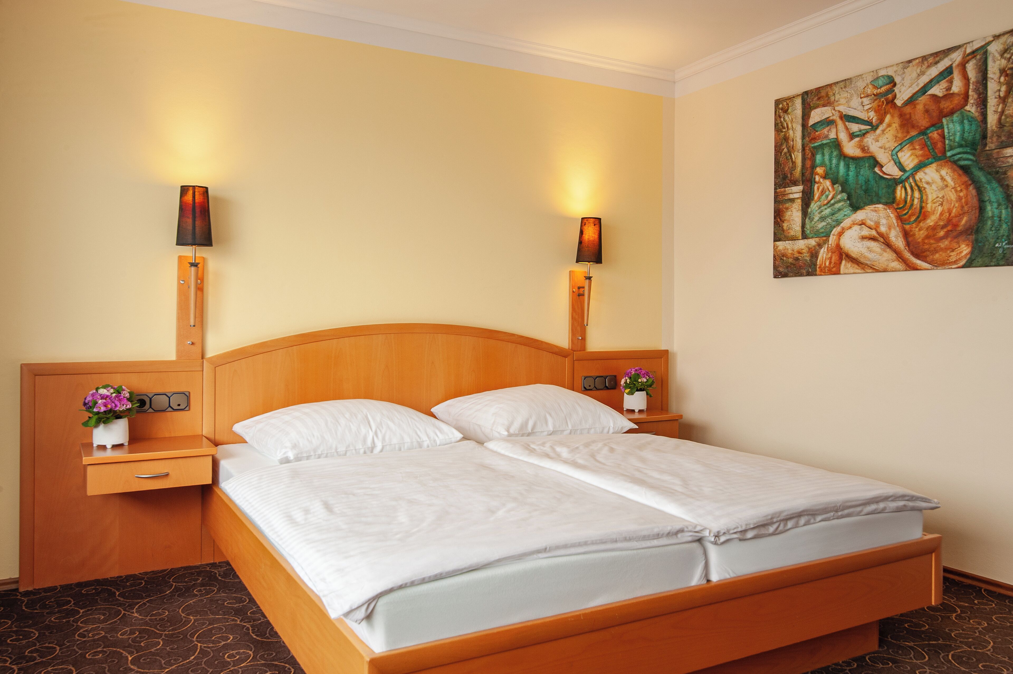Una cama doble con cabecera de madera, dos mesitas de noche con lámparas, una mesita con un jarrón de flores y un cuadro en la pared.