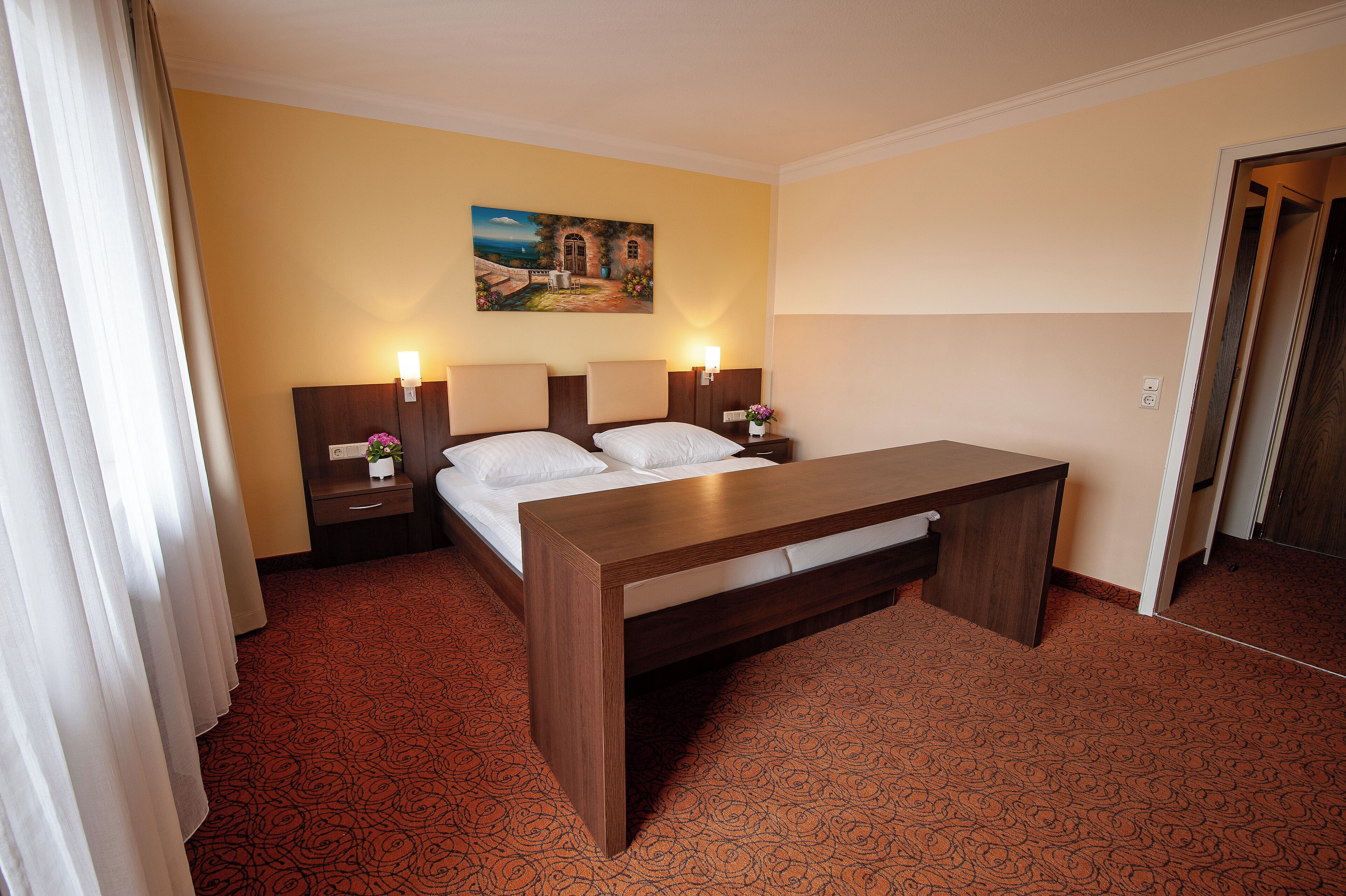 A hotel room with a bed, bedside tables, a wooden desk, a painting on the wall, and a patterned carpet.