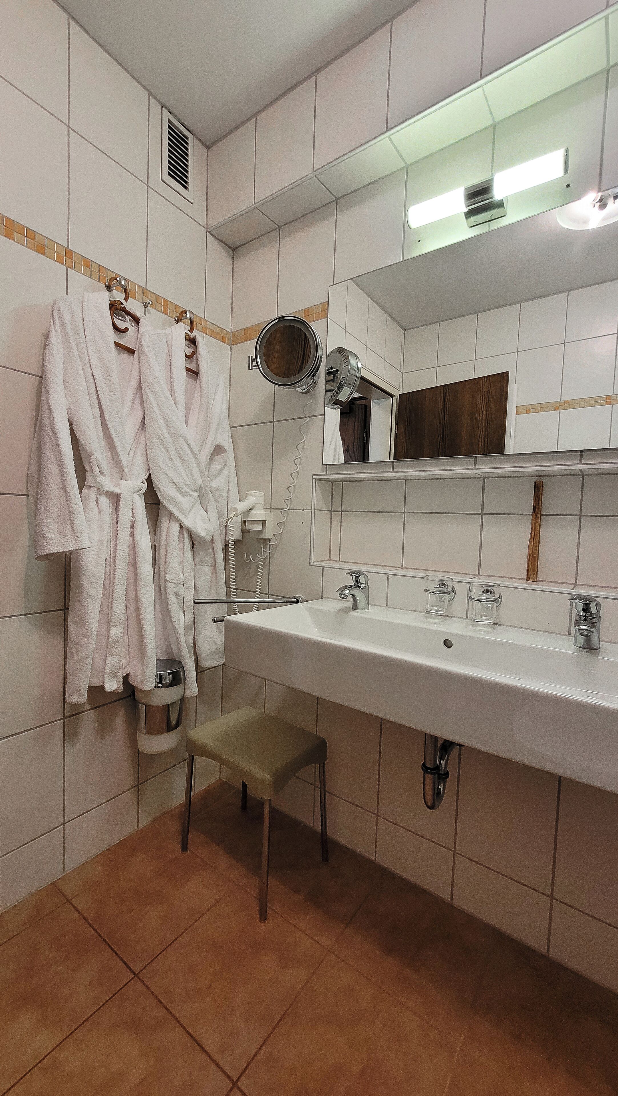 Une salle de bain avec un lavabo blanc, deux miroirs, un porte-serviettes avec deux peignoirs blancs, un petit tabouret et une bouteille de shampoing.