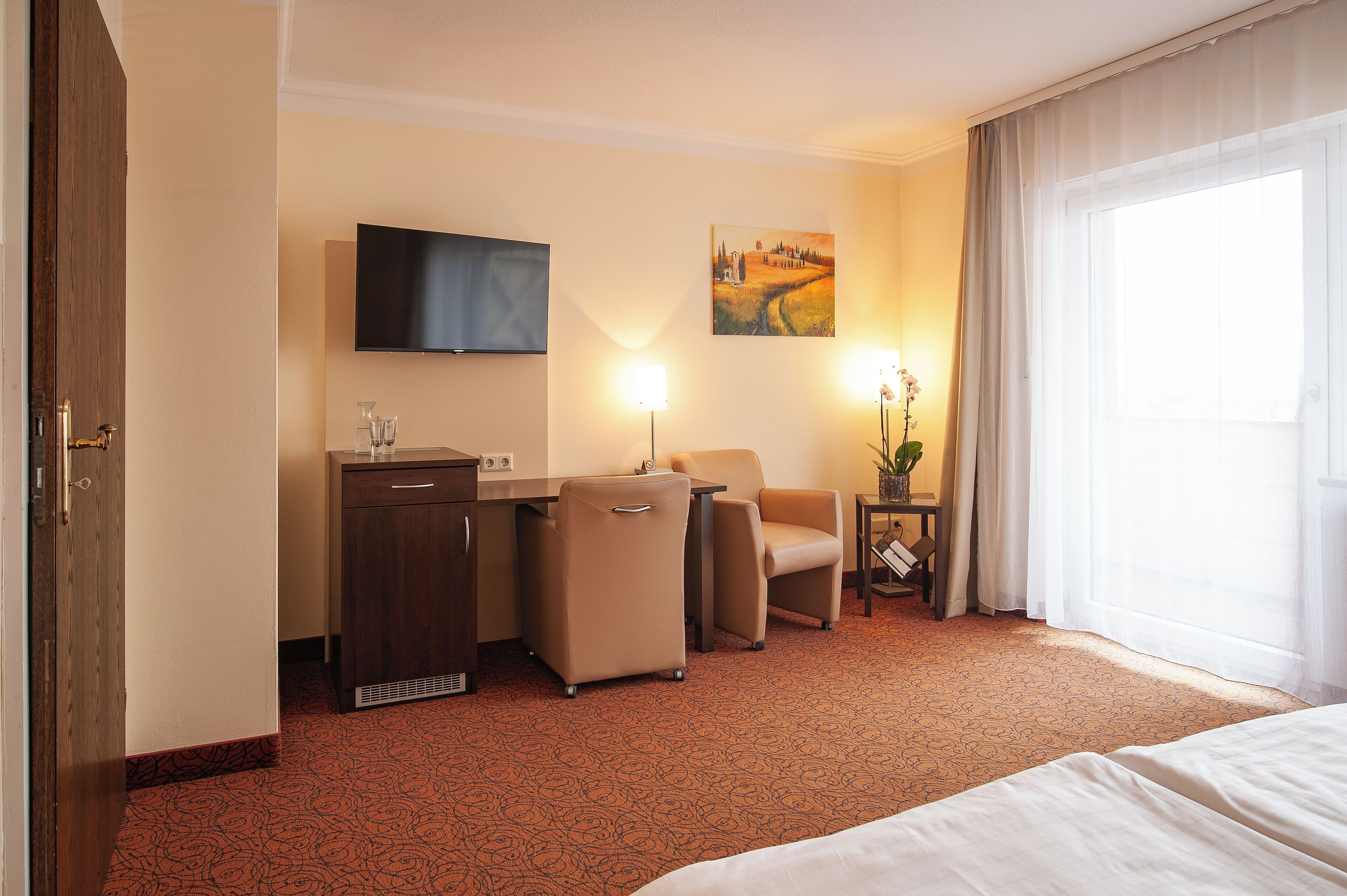 Une chambre d’hôtel avec un lit, un bureau, deux chaises, une télévision, un tableau et une fenêtre avec des rideaux.