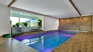 A modern indoor swimming pool with a wooden deck, stone wall, and large windows. - LuCA HOTEL (Bodenmais)