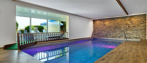 A modern indoor swimming pool with a wooden deck, stone wall, and large windows.