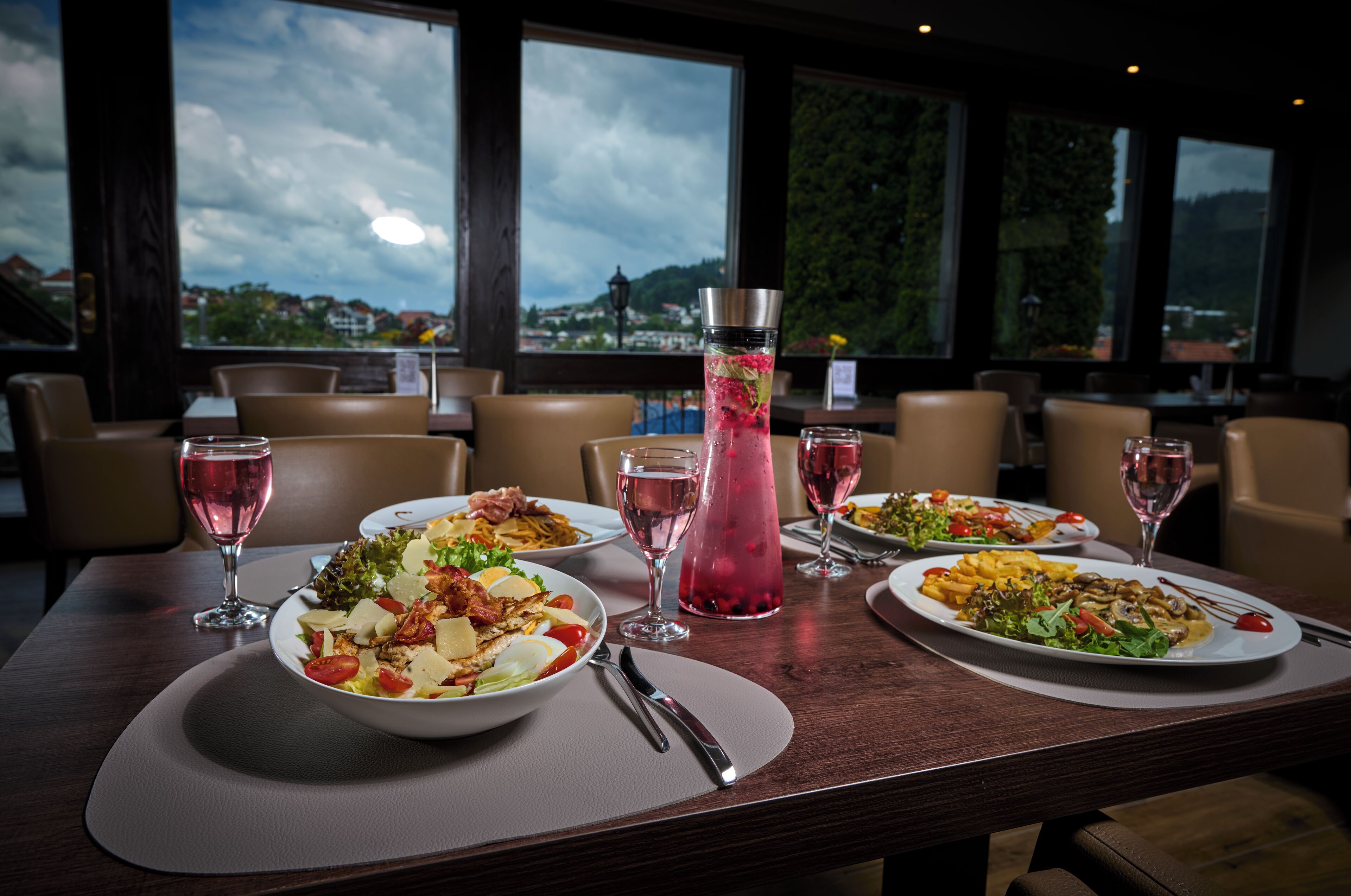 Una mesa de comedor con platos de comida, copas de vino y una bebida alta en una jarra metálica.