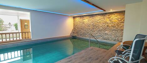 An indoor swimming pool with a wooden deck and chairs, a stone wall, and a window with a view.