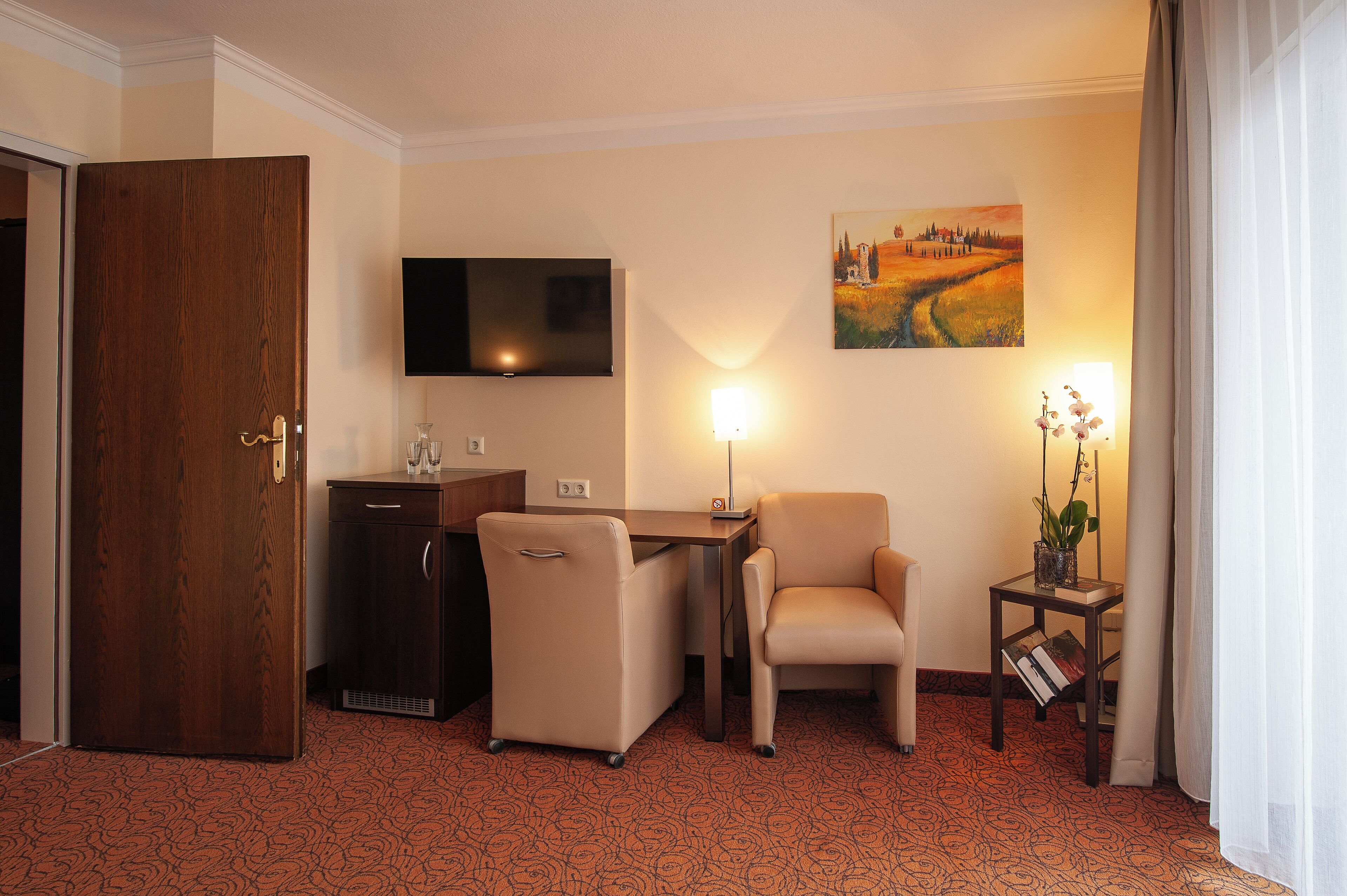 Habitación de hotel con puerta de madera, televisor montado en la pared, un cuadro, escritorio con silla, una mesita con una planta y suelo alfombrado.