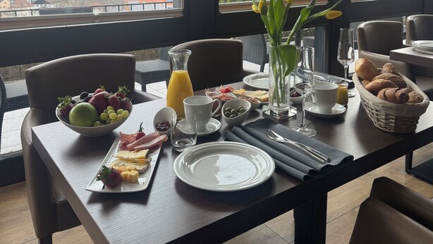 A breakfast table set with fruits, bread, and a glass of orange juice, overlooking a cityscape. - LuCA HOTEL (Bodenmais)