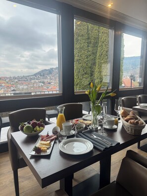 A breakfast table set with fruits, bread, and a glass of orange juice, overlooking a cityscape. - LuCA HOTEL (Bodenmais)