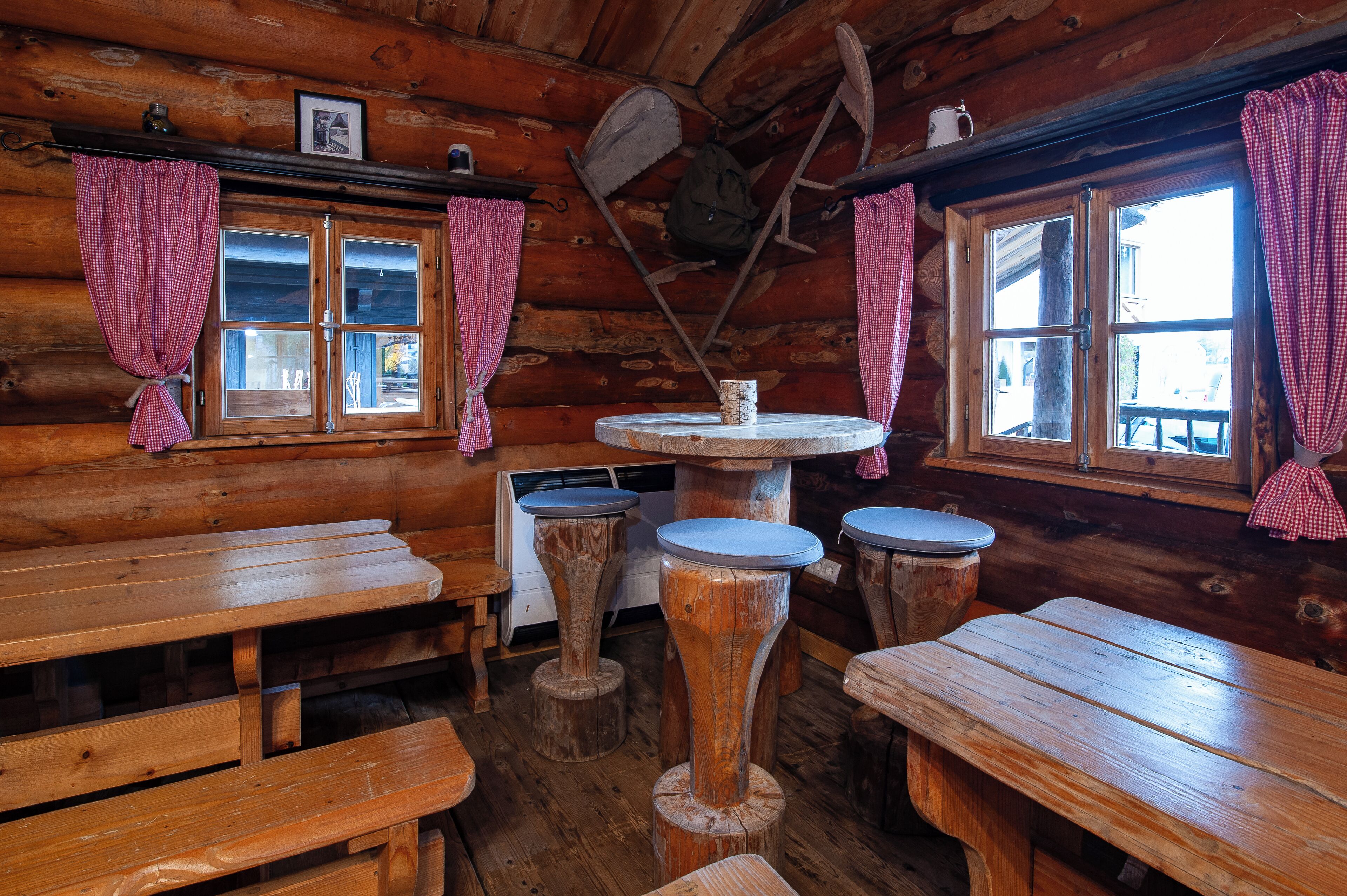 A rustic wooden cabin with wooden benches and tables, a central round table with stools, and checkered curtains on the windows.