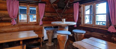 A rustic wooden cabin with wooden benches and tables, a central round table with stools, and checkered curtains on the windows.