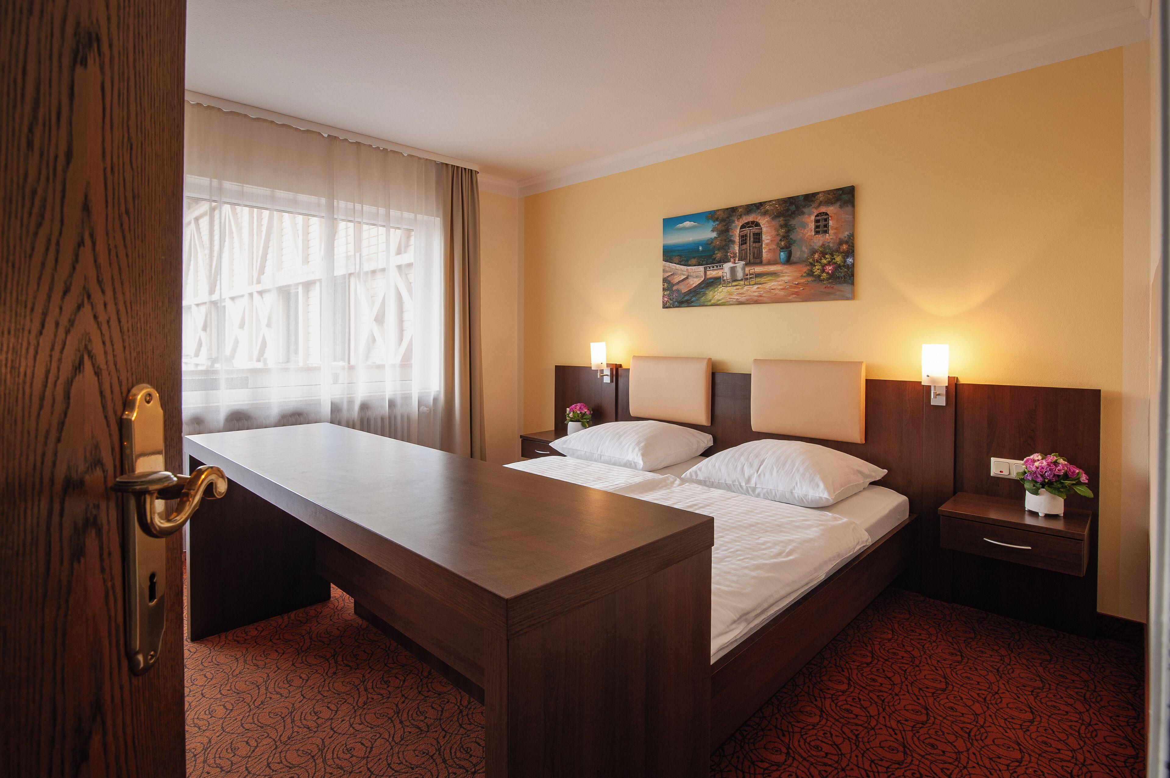 Ein Hotelzimmer mit Bett, Nachttischen, Schreibtisch, einem Bild an der Wand und einem Fenster mit Vorhängen.