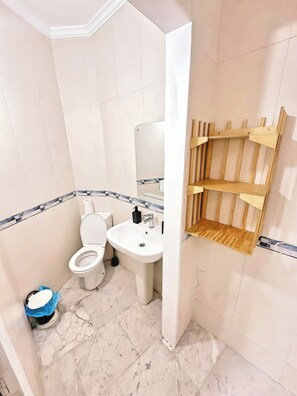 Et badeværelse med toilet, vask, spejl og en træhylde.