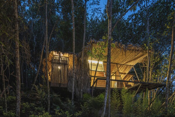 Una estructura tipo casa del árbol con techo de paja y vigas de madera, rodeada de un bosque denso.