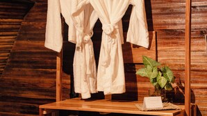 Un porte-peignoir en bois avec deux peignoirs blancs, un panier en osier, un ventilateur et une petite plante en pot.