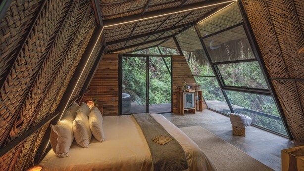 Une chambre à coucher confortable, de forme triangulaire, dotée d’une grande fenêtre qui offre une vue sur une végétation luxuriante.
