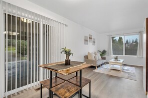 A modern living room with a wooden coffee table, a sofa, and a dining table. There are potted plants on the coffee table and a view of the outdoors through large windows. - Central Bellevue Retreat  (Bellevue)