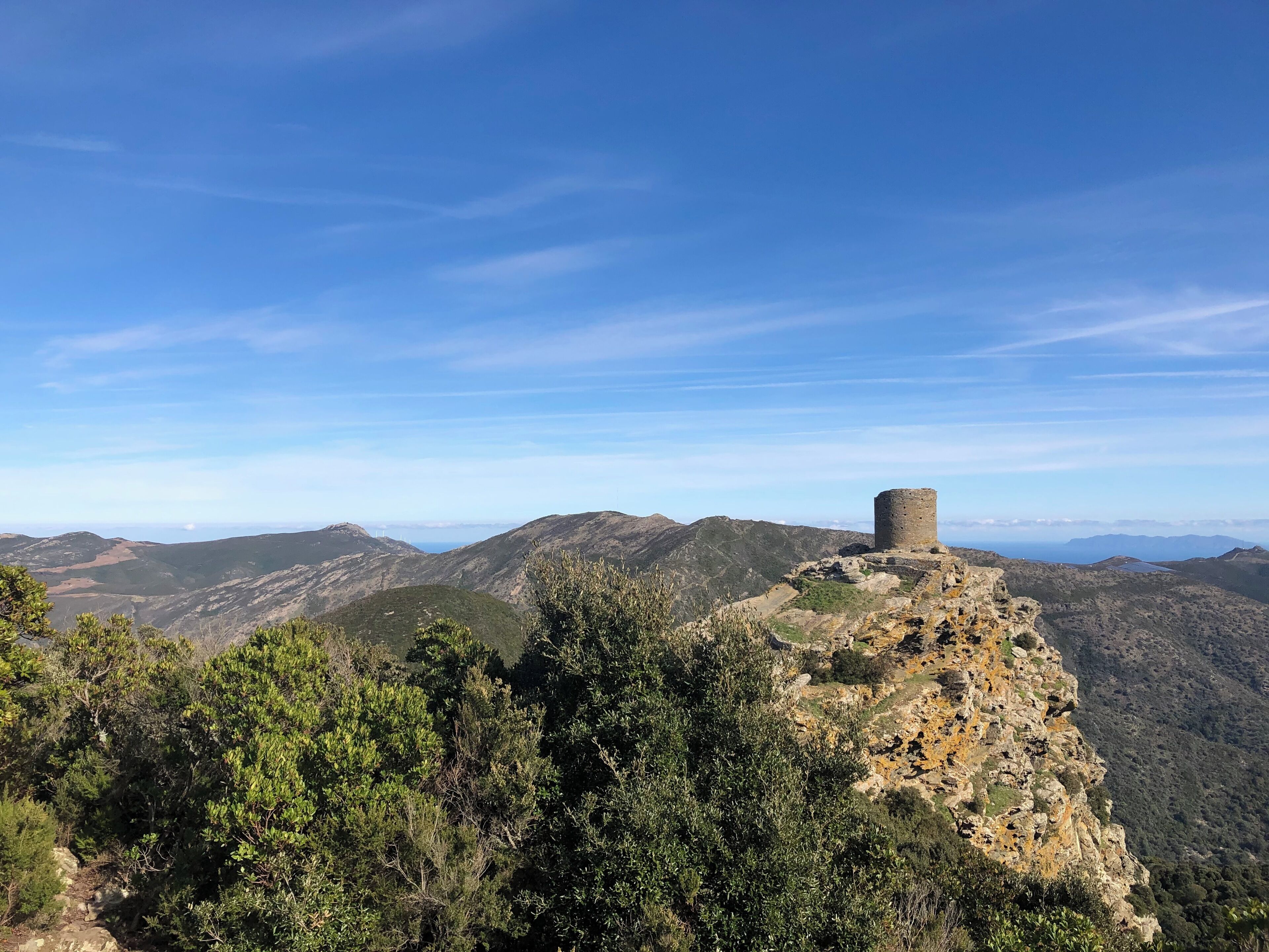 A historical stone tower on a mountain peak with surrounding forest and distant hills.